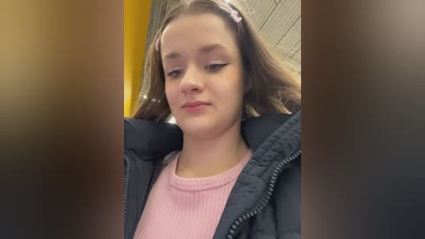 Video of a young girl with light skin, brown hair, and pink hair clips, wearing a pink sweater under a black jacket, in a blurred indoor setting with a yellow wall.