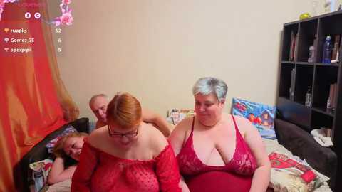 Video of a woman with short, light blue hair in a red lace bra, sitting on a bed with a man and a child, surrounded by toys and a TV screen showing a video feed.