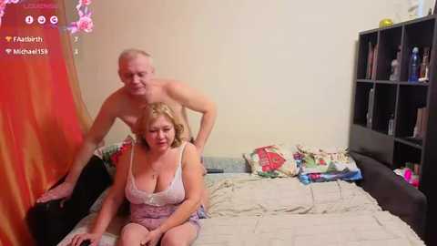 Video of a nude, muscular, bald man and a voluptuous, blonde woman in a bedroom, wearing a white lace bra and shorts.