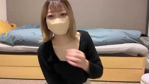 Video of an Asian woman with shoulder-length blonde hair, wearing a black long-sleeve shirt, beige mask, and beige gloves, standing in a minimalist bedroom with a blue bedspread and yellow bed frame.