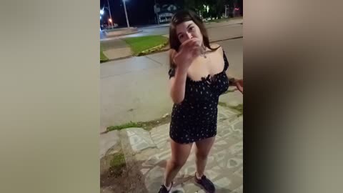 A video of a young woman with light skin and long brown hair, wearing a black polka-dotted dress, standing on a sidewalk at night.