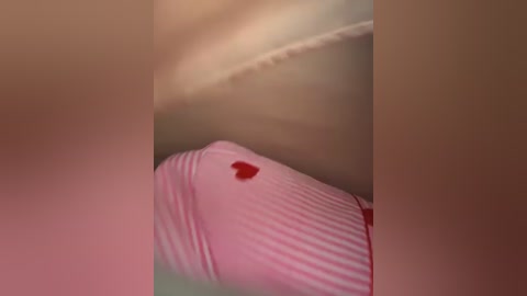 Video of a person lying on their side, wearing a pink striped shirt with a small red heart patch. The background is blurred, with beige and light brown tones.