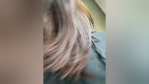 A blurry video of a person's shoulder and back, showing light hair and a blue shirt, with a green wall in the background. The image is out of focus.