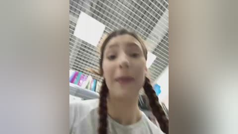 Video of a young woman with fair skin and long brown hair in braids, wearing a white shirt, taken from a low angle, indoors with a grid-patterned ceiling and colorful bookshelf in the background.