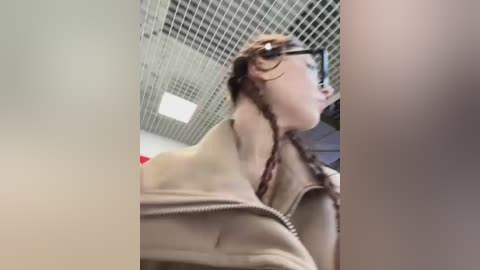 A video of a woman with glasses and braided hair, wearing a beige jacket, looking upward, in a room with a grid ceiling.