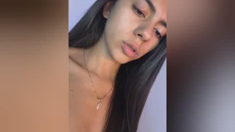 A close-up video of a young woman with long, straight black hair, light brown skin, and dark eyes, wearing a delicate necklace. The background is blurred, focusing on her face.
