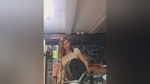 Video of a woman with shoulder-length brown hair in a beige t-shirt, using a weight machine in a dimly-lit gym with exposed concrete ceiling and metal racks in the background.