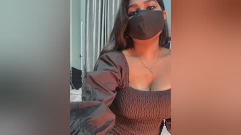 A video of a South Asian woman with long black hair, wearing a black face mask, a brown off-shoulder top, and a gold necklace, indoors.