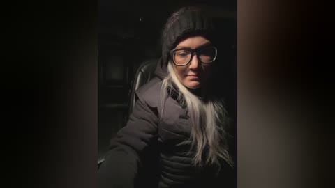 Video of a middle-aged woman with long, straight blonde hair, wearing glasses and a black knit hat, seated in a dimly lit car.