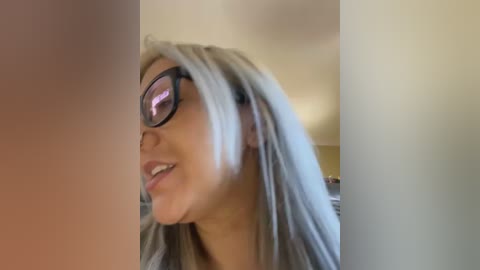 Video of a light-skinned woman with long, platinum blonde hair and large, black-rimmed glasses, smiling. The background is a blurred, beige-colored room with a dim light source.