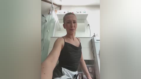 A video of a fair-skinned woman with a ponytail, wearing a black tank top and gray apron, standing in a kitchen with white cabinets and a glass door.