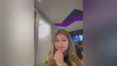 Media: A young woman with long, wavy blonde hair and fair skin stands in a modern, dimly-lit room with purple LED lighting, her finger to her lips in a \"shh\" gesture.