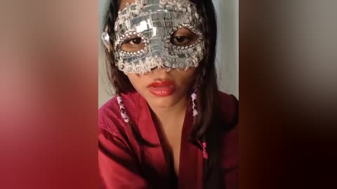 A video of a young Asian girl with straight black hair and light skin, wearing a silver lace masquerade mask and a burgundy robe.