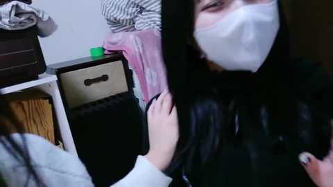 Video of a young woman with long black hair wearing a white mask and a black coat, assisting another person with a pink scarf, in a cluttered, dimly-lit room.