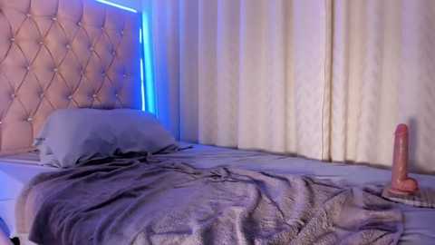 Video of a dimly lit bedroom with a tufted headboard, blue lighting, a gray blanket, and a large, realistic sex toy on the bed.