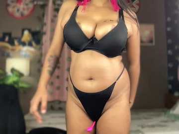 Video of a curvy woman with medium brown skin, wearing a black lace bra and thong, revealing ample cleavage and tattoos on her arms, standing indoors with a blurred background.