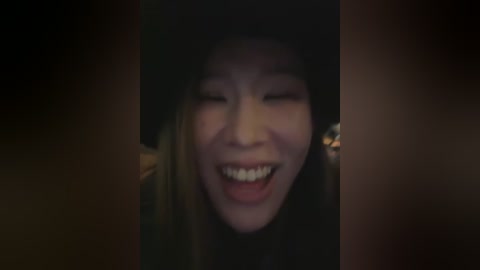 A close-up video of a smiling Asian woman with fair skin, wearing a black hat and a dark coat, laughing joyfully. The background is dark and blurry, emphasizing her expressive face.