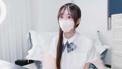 Video of an Asian woman with long, straight brown hair wearing a white mask, plaid tie, and white blouse, sitting on a white bed in a minimalist, bright room with white walls and a blue cabinet.