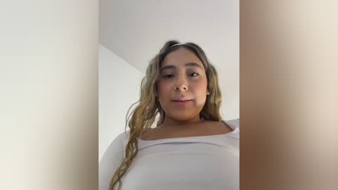 Video of a young Latina woman with light brown skin, long wavy hair, and a confident expression. She wears a white long-sleeved top, standing in a simple indoor setting with white walls.