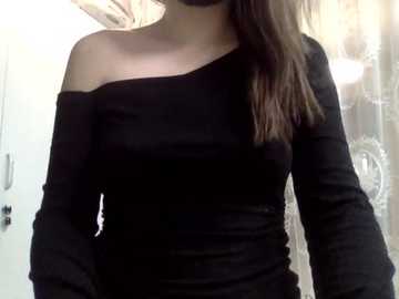 Video of a young woman with fair skin and long brown hair, wearing a black off-the-shoulder long-sleeve dress, standing in a bathroom with a white door and glass shower enclosure in the background.