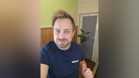 A video of a young man with short, spiky hair, light skin, and a trimmed beard, giving a thumbs-up. He wears a navy t-shirt with \"blog\" in white. Background includes a green wall, wooden paneling, and a potted plant.
