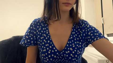 A video of a young woman with medium-length dark hair, wearing a blue V-neck dress with white polka dots, sitting on a dark couch in a dimly lit room with beige walls.