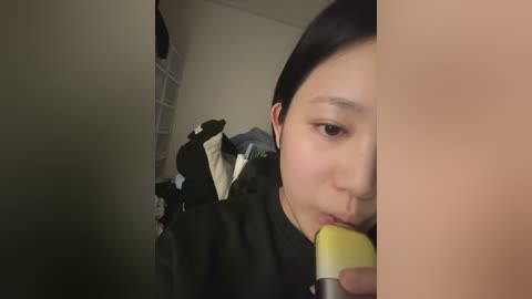 A video of an Asian woman with fair skin and straight black hair, wearing a black dress, using a yellow and white hairbrush to comb her hair. She is indoors with white-framed windows in the background.