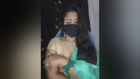 Video of a South Asian woman with medium-dark skin, long black hair, wearing a green saree, black mask, and gold jewelry, seated in a dimly lit room with greenery in the background.