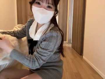 Video of an Asian woman with long dark hair, wearing a face mask and a grey knit cardigan, sitting on a wooden floor in a dimly lit room with beige walls.