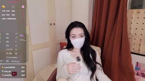 Video of a young woman with long black hair, wearing a white face mask and a white cardigan, sitting on a bed in a cozy, beige-toned room with a pink blanket and brown curtains.