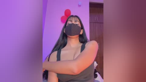 Video of a young woman with long black hair, wearing a black face mask, black tank top, and a red heart balloon, in a dimly lit room.