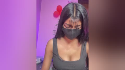 Video of a young woman with long black hair, wearing a black face mask and a sleeveless gray top, indoors with purple lighting, red balloons, and minimalistic wall art in the background.