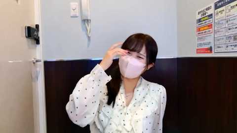 Video of a woman in a white blouse with black dots, wearing a pink surgical mask, adjusting her hair in a medical office with a white door and a bulletin board with news articles on the wall.