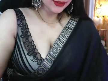 Video of a South Asian woman with fair skin, wearing a black saree with a grey, floral-patterned blouse. She has long, straight black hair and is adorned with large, ornate earrings and red lipstick.