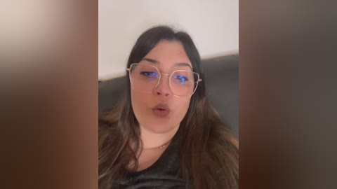 Video of a young woman with long, dark hair and light skin, wearing glasses and a black top, blowing a kiss.