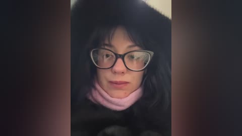 Video of a woman with light skin and straight black hair, wearing black-framed glasses and a black fur hat, set against a dark background, with a subtle pink scarf visible.
