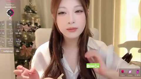 Video of an Asian woman with long brown hair, light skin, and a slim physique, wearing a white blouse. She's holding a phone, with a calendar and a green \"Dana\" logo visible. Background shows a Christmas tree and a lit candle.