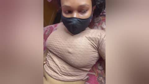 Video of a Black woman with medium-brown skin and curly hair, wearing a black face mask, beige top, and floral-patterned skirt, seated indoors with blurred background.