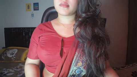 Video of a South Asian woman with long, wavy dark hair, wearing a red top and a patterned shawl, seated in a bedroom with a bed, a black headboard, and a yellow wall clock.