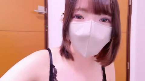 Video of an East Asian woman with shoulder-length brown hair, wearing a white face mask and black lace-trimmed bra. She has fair skin and is indoors, standing near a wooden door.