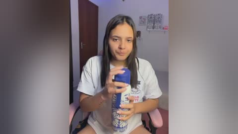Video of a young Latina woman with long black hair, wearing a white t-shirt and shorts, sitting on a pink chair, holding a blue water bottle, in a cozy, dimly-lit room with white walls and a small shelf.