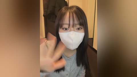 Video of a young Asian woman with long black hair, wearing a white surgical mask, gray sweater, and a gray scarf, in a dimly lit room with beige walls.