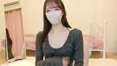 Video of an Asian woman with long black hair, wearing a gray ruffled top and white face mask, seated on a white bed in a beige room with a pink curtain and white furniture.