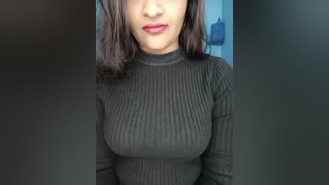 Video of a South Asian woman with medium-brown skin, medium-length black hair, wearing a ribbed black long-sleeve top, standing in front of a blurred blue wall, showing her upper body from chest to just below the nose.