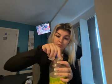 Video of a young woman with long blonde hair, wearing a black top, holding a yellow drink and making a peace sign. Background shows a TV on a blue wall and a white door.