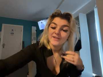 Video of a young woman with blonde hair, wearing a black top, adjusting her hair in a bedroom with teal walls and a white door.