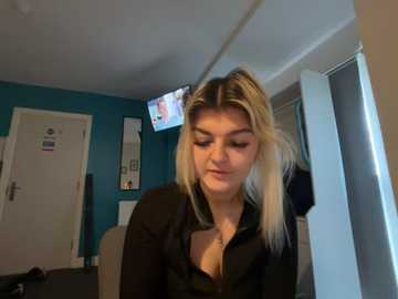 Video of a blonde woman with straight hair, wearing a black blouse, sitting in a modern room with blue walls, white door, and a TV mounted on the wall.