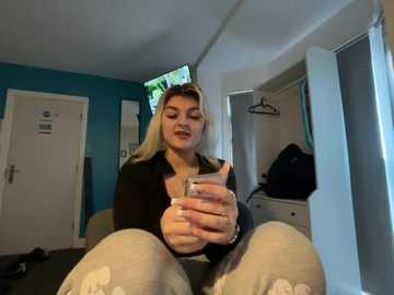 Video of a blonde woman with fair skin, wearing a black top and light pants, sitting on a bed, holding a smartphone, in a blue-painted room with a TV, mirror, and clothes hanging.