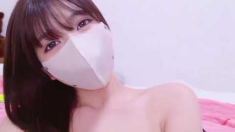 Video of a young Asian woman with straight black hair and fair skin, wearing a white mask and pink top, lying on a pink bed in a bright room.