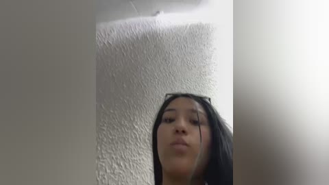 A video of a young woman with medium skin tone, long black hair, and light makeup, making a pouty face, captured from a low angle. She stands in a room with a textured white ceiling and a bright light source on the right.